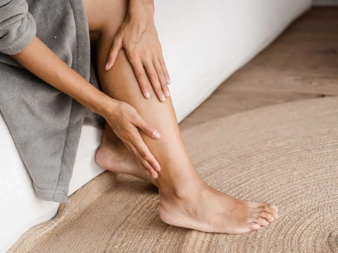 Une personne se tient la jambe gauche, peut-être à cause d'une douleur jambe gauche. Elle est assise sur un lit, enveloppée dans un peignoir gris.