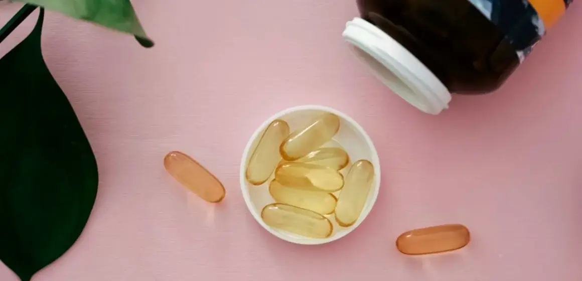 Gélules de collagène et vitamine C sur fond rose, à côté d'une feuille verte.