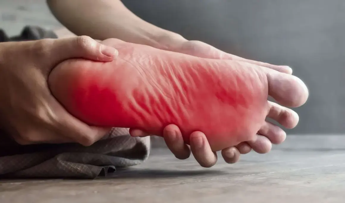 Main gauche, la plante du pied est tenue et massée, avec une zone rouge vif indiquant une douleur au pied gauche.