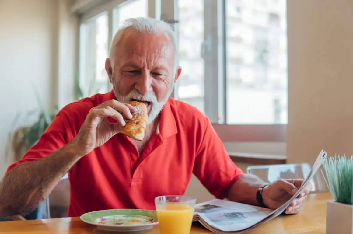 Un homme âgé mange un croissant en lisant le journal, un verre de jus d'orange à côté. Une scène qui illustre les besoins nutritionnels de la personne âgée.