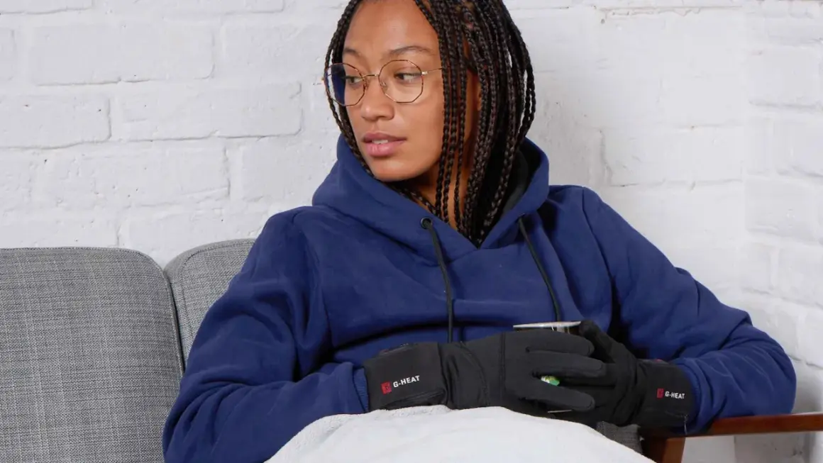 Femme avec des tresses, lunettes, sweat bleu et gants chauffants G-HEAT, assise sur un canapé, elle semble avoir toujours froid.