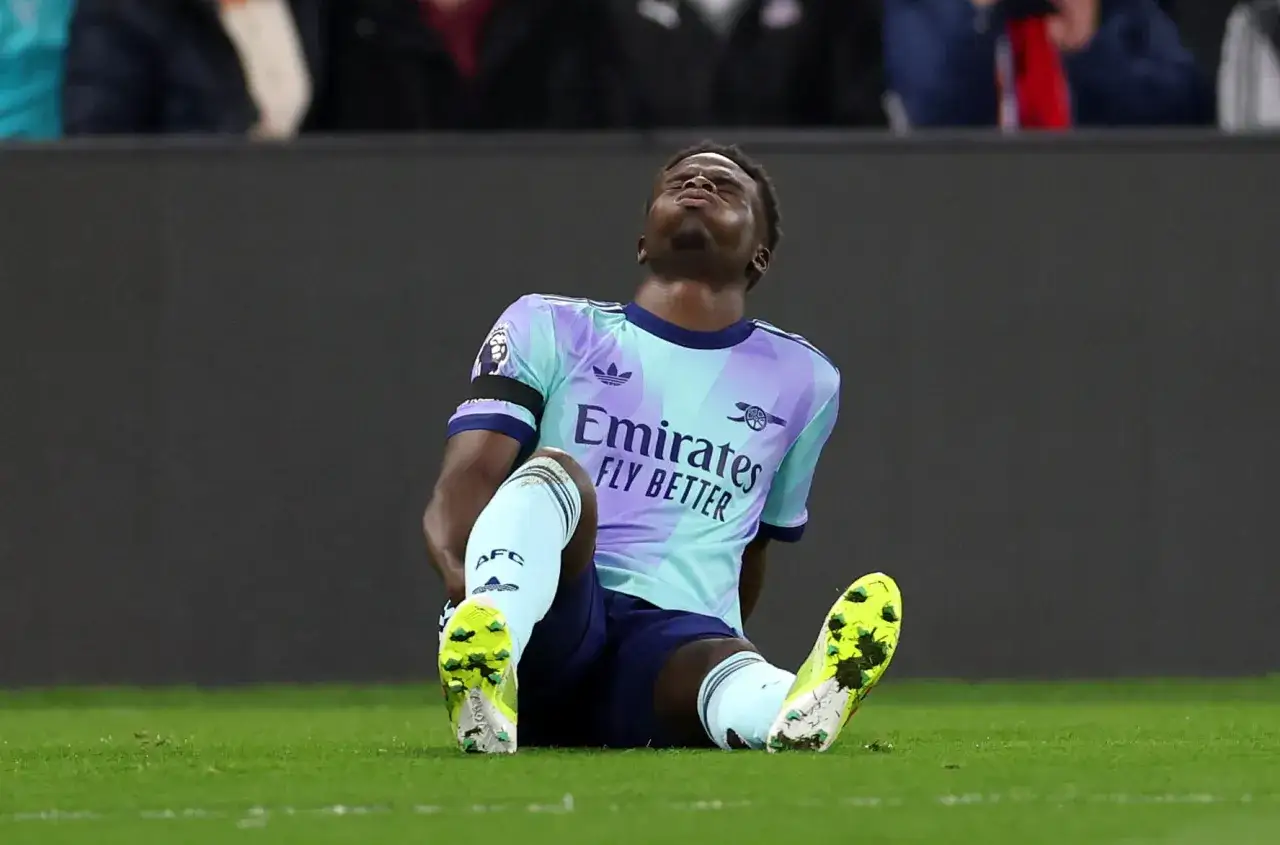 Un joueur de foot, le visage tourné vers le ciel, s'assoit sur la pelouse, une douleur visible à la jambe. Sa pointe derrière la cuisse semble indiquer un problème.