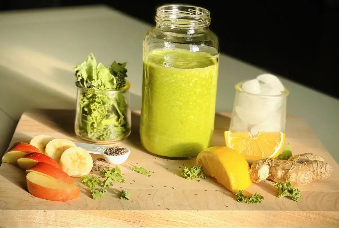 Un verre de smoothie vert, des tranches de pomme, de banane, du gingembre, du citron et du chou frisé, ingrédients parfaits pour une cure detox maison.