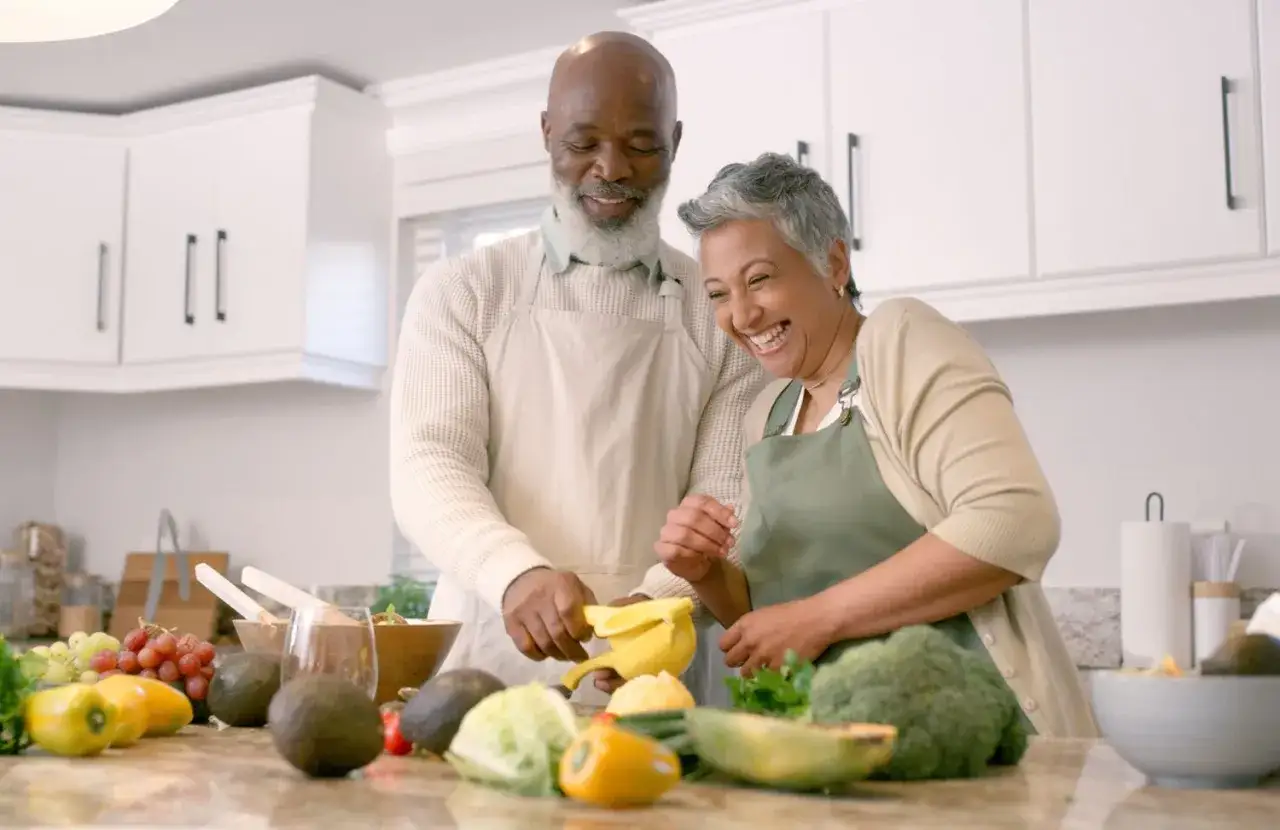 Couple âgé préparant des fruits et légumes frais, une scène joyeuse qui illustre les besoins nutritionnels de la personne âgée.