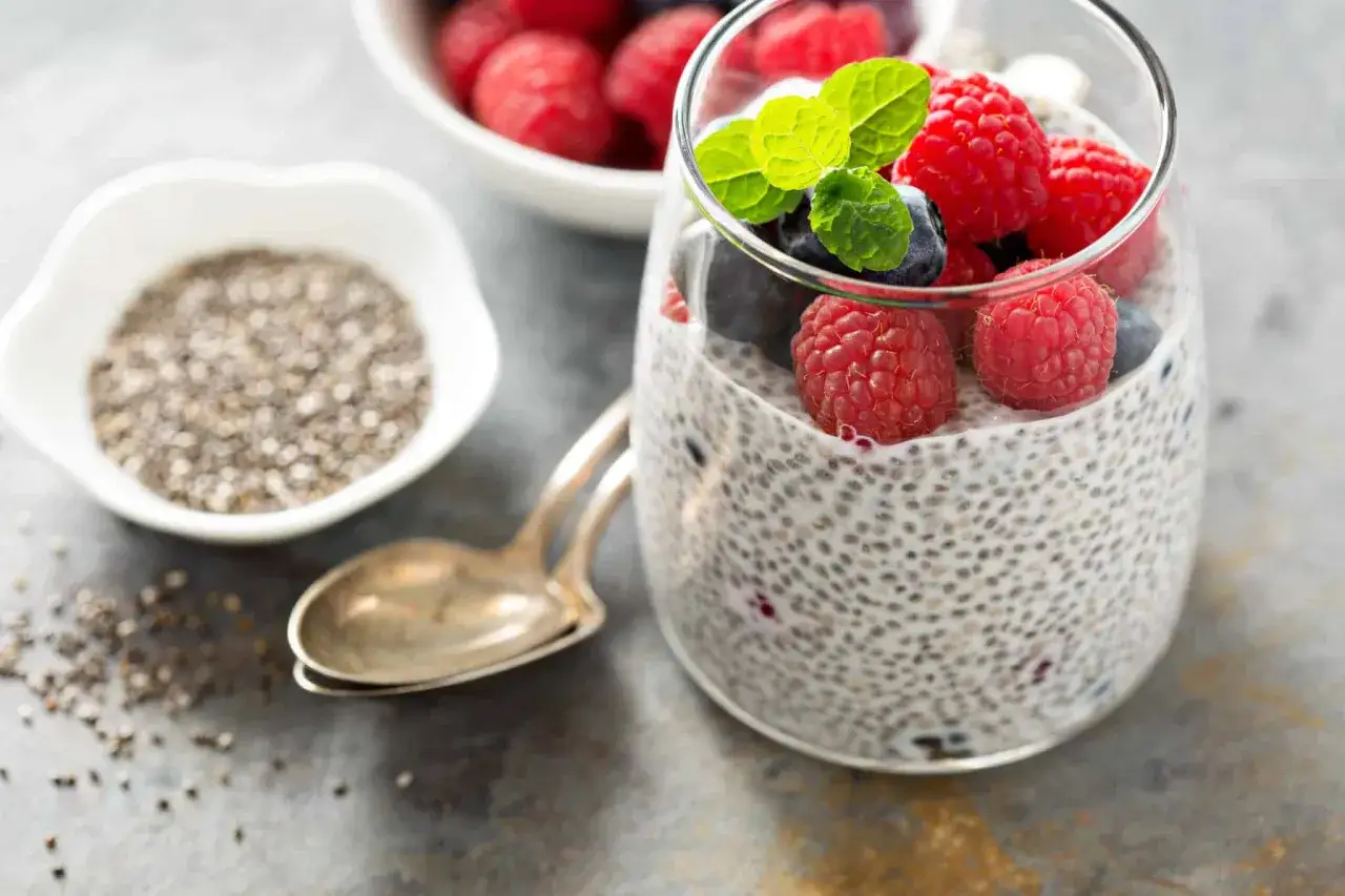 Un verre de pudding aux graines de chia, garni de framboises, myrtilles et menthe. À côté, un bol de graines de chia et une cuillère.