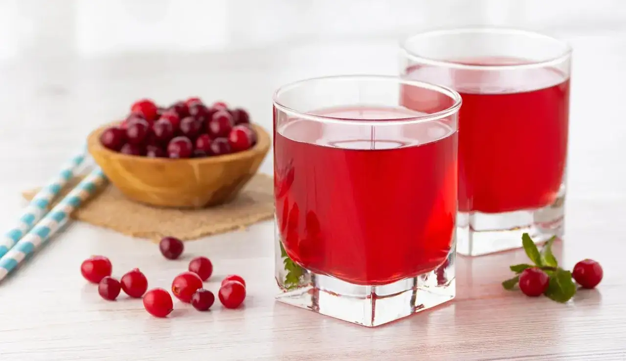 Deux verres de jus de cranberry, une boisson réputée pour aider à prévenir les infections urinaires. Des baies fraîches et une petite branche de menthe complètent la scène.