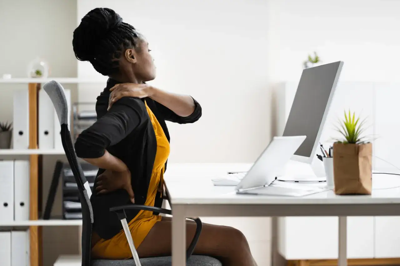 Femme assise à un bureau, se tenant le cou et le bas du dos, souffrant de muscles du cou douloureux après une longue journée de travail.