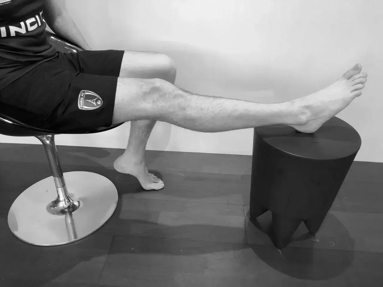 Homme en short de sport, jambe tendue, pied sur un tabouret. Rééducation après blessure au ligament croisé du foot.