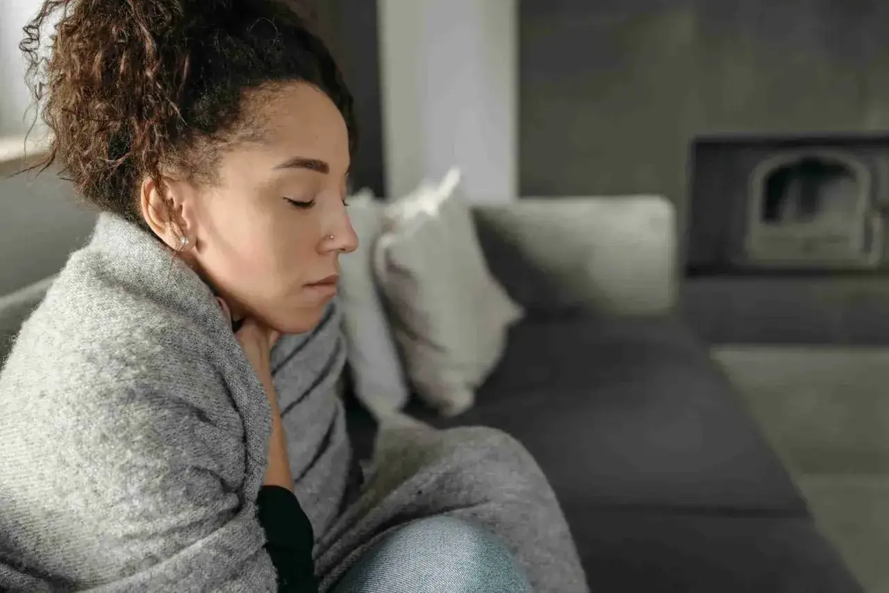 Une femme emmitouflée dans une couverture grise, les yeux fermés, médite sur pourquoi j'ai toujours froid. Le canapé gris et le foyer flou créent une ambiance introspective.