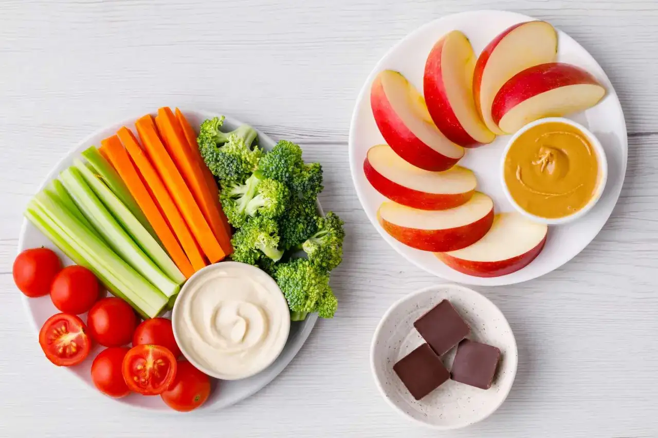 Assiette de légumes croquants (carottes, céleri, brocolis, tomates) avec houmous, pommes tranchées avec beurre de cacahuète, et carrés de chocolat. Un délice à faible indice glycémique.