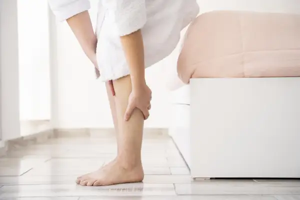 Une personne en peignoir blanc se tient près d'un lit, se massant la jambe. Elle cherche des remèdes de grand-mère pour ses jambes lourdes.