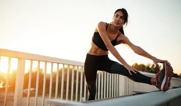 Femme s'étirant les ischio-jambiers, une conséquence d'un entraînement intense. Le soleil se lève sur un paysage flou.