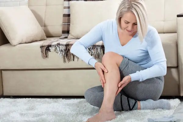 Femme assise sur un tapis blanc, touchant sa jambe. Elle ressent une douleur derrière la cuisse sans effort, visiblement préoccupée.
