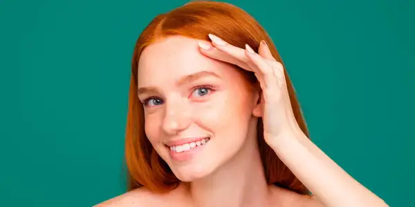 Une jeune femme rousse souriante, peau parsemée de taches de rousseur, touche son front. Elle utilise peut-être un soin au zinc pour la peau.