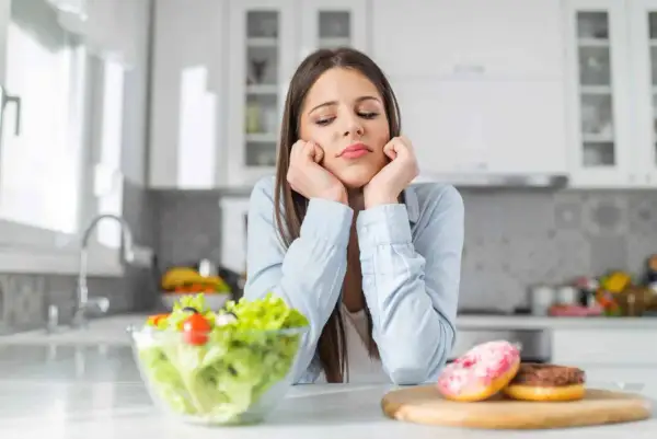 Une jeune femme dépitée face à une salade et des donuts, symbolisant le dilemme des aliments à éviter pour la dermite séborrhéique.