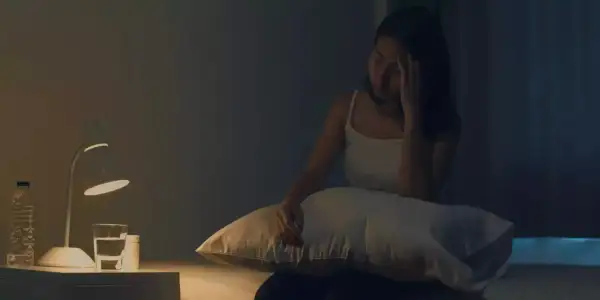 Femme insomniaque, le front serré, envisageant une cure de magnésium. Sur la table de nuit, une lampe, une bouteille d'eau et des médicaments.