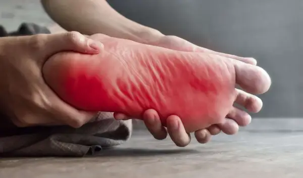 Main gauche, la plante du pied est tenue et massée, avec une zone rouge vif indiquant une douleur au pied gauche.