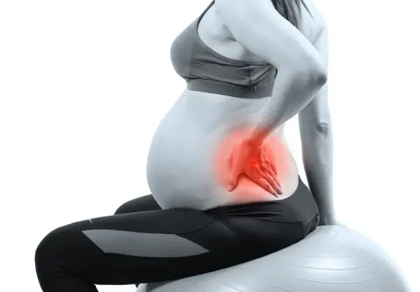 Femme enceinte assise sur un ballon de gym, main sur le bas du dos, zone rouge indiquant une douleur au bassin pendant la grossesse.