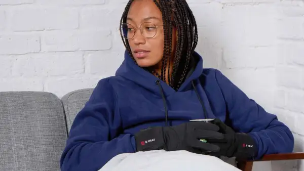 Femme avec des tresses, lunettes, sweat bleu et gants chauffants G-HEAT, assise sur un canapé, elle semble avoir toujours froid.
